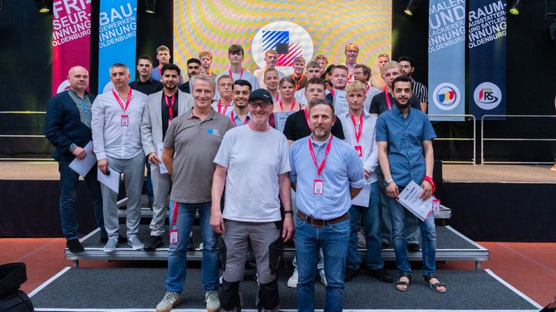 Großes Gruppenfoto von Lehrlingen und Handwerksgesellen auf einer Bühne während einer Innungs- und Abschlussveranstaltung in Oldenburg. Die Teilnehmenden stehen gestaffelt vor einer beleuchteten Leinwand, viele halten Zertifikate; im Hintergrund sind bunte Banner mit Aufschriften verschiedener Innungen (z. B. Friseur-, Bau- und Malerinnung). Das Bild vermittelt feierliche Stimmung und Gemeinschaft im Handwerk.
