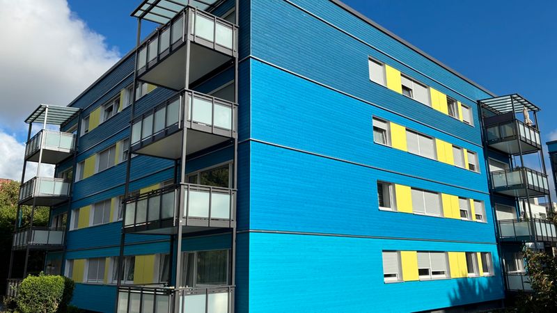 Foto eines modernen, blau gestrichenen Mehrfamilienhauses mit mehreren Glasbalkonen und gelben Fensterakzenten, aufgenommen bei klarem, sonnigem Himmel. Vordergrund mit kleiner Beetfläche und Gehweg, klare Linien der Fassade sowie Kontrast zwischen Blau und Gelb prägen das Bild.
