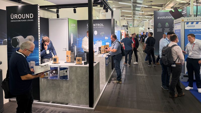 Hauptmotiv: Ein Messestand in einer Messehalle mit ausgestellten Produkten und mehreren Besuchern, die sich informieren und austauschen. Im Vordergrund ist ein Stand mit Produktpräsentationen und Informationsmaterial, im Hintergrund weitere Stände und Laufwege der Messe.