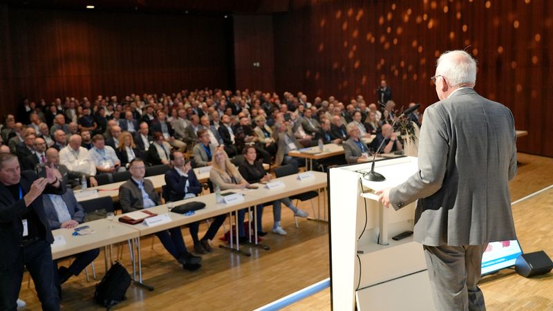 Ein Redner am Podium, der mit Mikrofon vor einem großen, sitzenden Publikum in einem Konferenzsaal spricht.