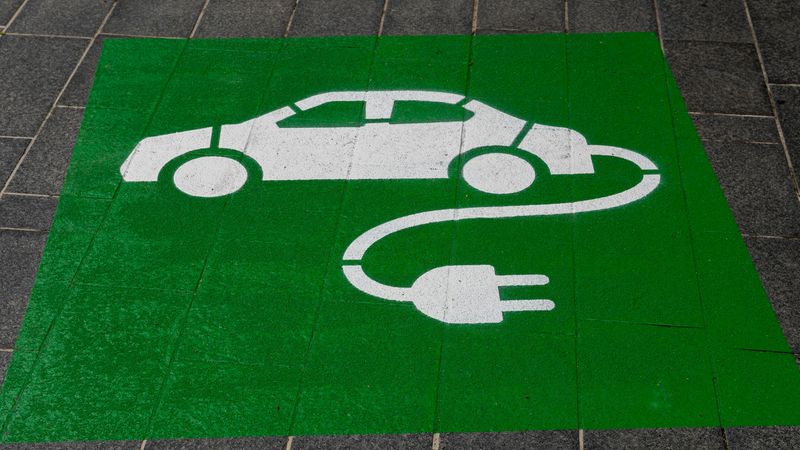 Weißes Piktogramm eines Elektroautos mit angehängtem Ladekabel und Stecker auf einem grünen Quadrat, auf Gehwegplatten gemalt — Hauptmotiv: Elektroauto-Ladezeichen auf Parkplatzmarkierung.