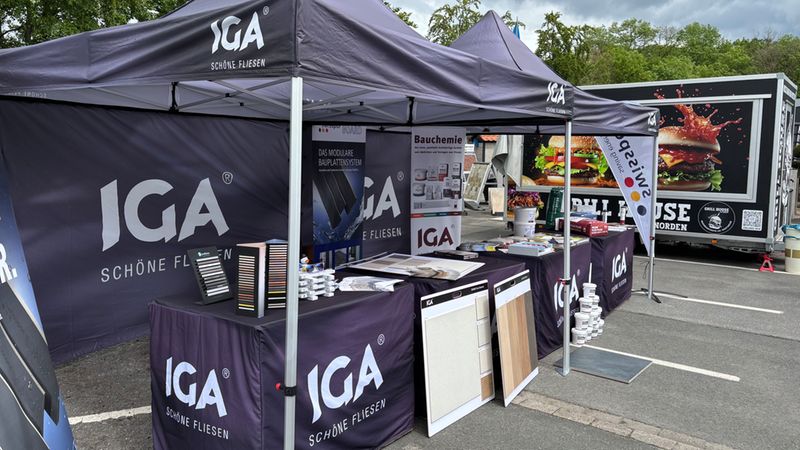 Outdoor-Messestand der Marke IGA mit dunklem Marken-Pavillon, mehreren Tischen mit Fliesenmustern, Musterplatten und Prospekten auf einem Parkplatz. Im Hintergrund ist ein Werbeanhänger mit Foodtruck-Motiv zu sehen; der Stand präsentiert Produktproben, Bauchemie-Informationen und Broschüren für Besucher.