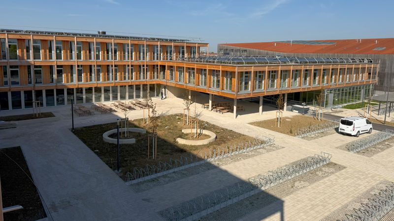 Weitwinkelaufnahme des Innenhofs eines modernen Holzgebäudes mit markanter Holzfassade und großen Fenstern. Der gepflasterte Hof zeigt Fahrradständer, Sitzbereiche, junge Bäume und einen Parkplatz mit einem weißen Transporter; auf den Dächern sind Solarpaneele zu sehen. Klare Tageslichtstimmung unter blauem Himmel.