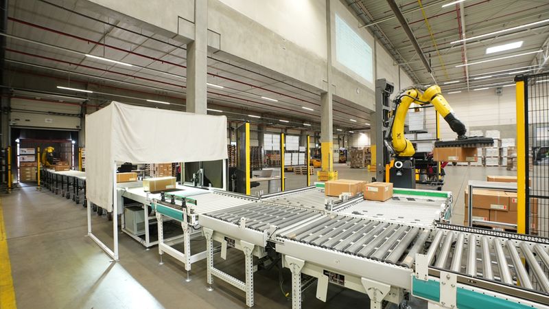 Hauptmotiv: automatisierte Förderanlage mit gelbem Industrieroboter und mehreren Paketen in einer großen Lagerhalle. Zu sehen sind Rollbahnen, Förderbänder, Kartons und eine Roboterzelle, die Pakete sortiert; helle Industriebeleuchtung und hohe Hallendecke betonen das Logistik- und Automatisierungsumfeld.