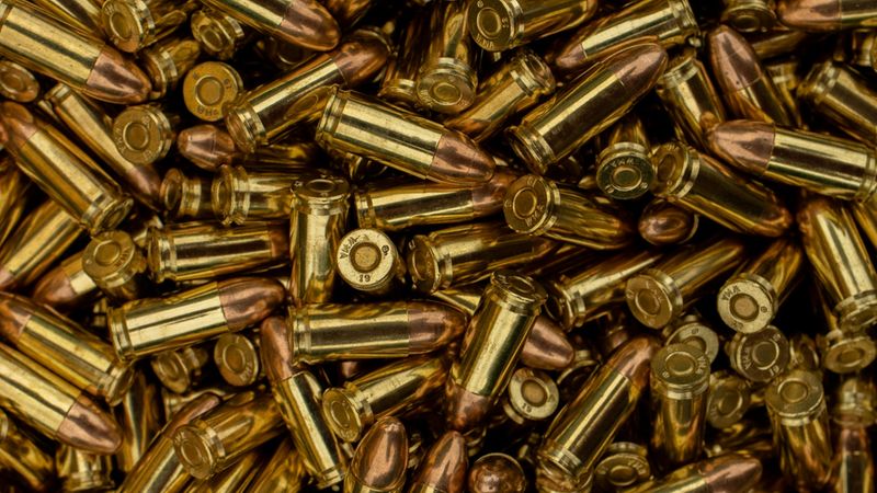 Nahaufnahme eines dichten Haufens goldener Patronen (Handwaffenmunition) mit glänzenden Messinghülsen und kupferfarbenen Geschossenden als zentrales Hauptmotiv.