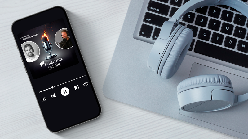 Nahaufnahme eines Smartphones, das den Podcast-Player der Sendung "FeuerTrutz ON AIR" mit Mikrofon-Icon und Sprecherporträts anzeigt. Daneben liegen weiße Kopfhörer auf einem silbernen Laptop auf hellem Schreibtisch. Modernes Audio- und Streaming-Setting, ideal für Podcasts und Medienproduktion.