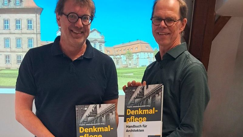 Hauptmotiv: das Fachbuch "Denkmalpflege". Zwei lächelnde Personen halten jeweils ein Exemplar des Buches "Denkmalpflege - Handbuch für Architekten" vor einem projizierten Architekturhintergrund. Die Szene wirkt wie eine Vorstellung oder Präsentation des Buches in einem Innenraum und betont Publikation, Denkmalpflege und Architektur.