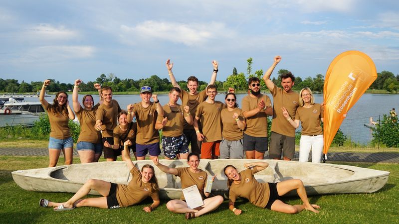 Hauptmotiv: Eine fröhliche Gruppe junger Erwachsener posiert gemeinsam vor einem Kanu am Ufer eines Sees. Die Personen tragen einheitliche braune T‑Shirts und feiern, einige stehen, andere sitzen oder liegen vor dem Kanu; im Hintergrund sind Wasser, Boote und eine orange Fahne zu sehen. Die Stimmung ist sommerlich und gemeinschaftlich.