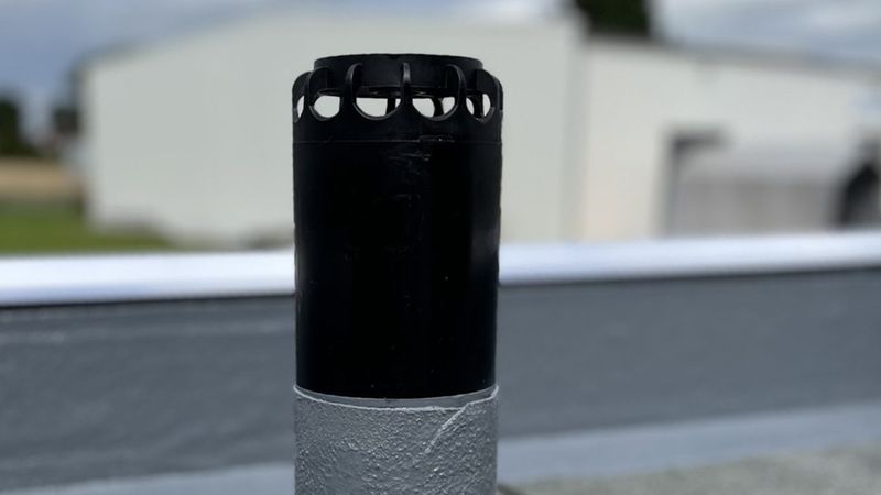 Nahaufnahme eines Dachentlüftungsrohrs (Hauptmotiv) mit schwarzer Belüftungskappe und grau abgedichteter Basis auf einem Flachdach, mit unscharfem Hintergrund.