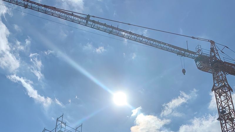 Ein großer Baukran ragt in den klaren blauen Himmel, wobei die helle Sonne direkt im Hintergrund scheint und einen leichten Lichteffekt erzeugt. Am unteren Bildrand sind zudem Teile eines Baugerüsts erkennbar.