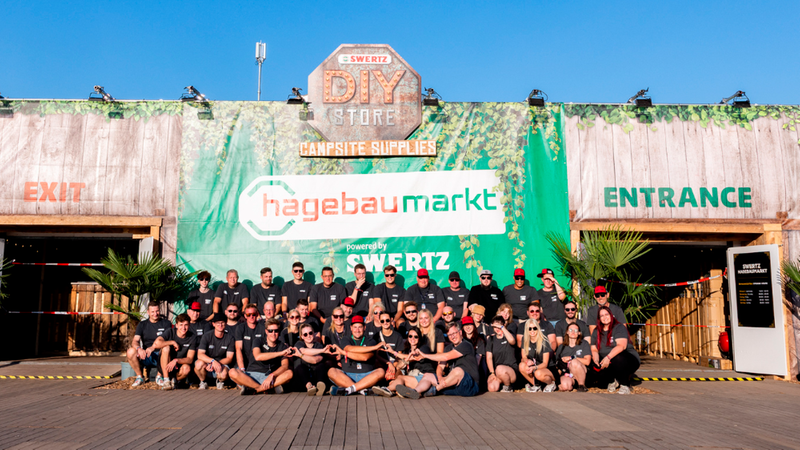 Eine große Gruppe von Menschen posiert fröhlich für ein Gruppenfoto vor der auffälligen Fassade des Swertz hagebaumarkt DIY Store, der als Campsite Supplies Shop gekennzeichnet ist.