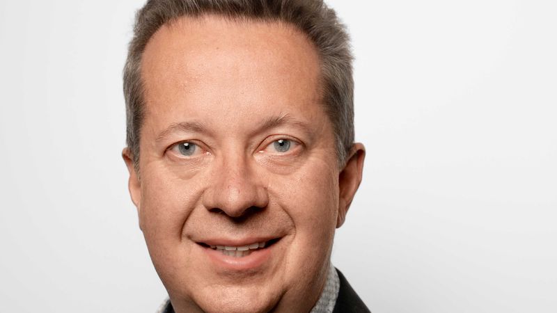 Ein professionelles Portrait eines freundlich lächelnden Mannes mittleren Alters, der ein elegantes Sakko und ein gemustertes Hemd vor einem neutralen, hellen Hintergrund trägt.
