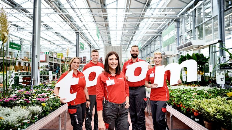 Ein freundliches Team von toom Baumarkt-Mitarbeitern in roter Arbeitskleidung steht in einem Gartencenter und hält gemeinsam große Buchstaben, die das Wort 'toom' bilden.
