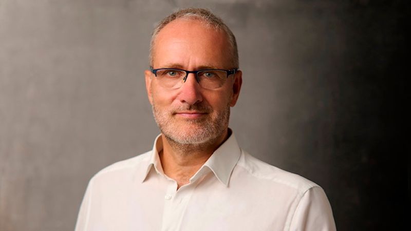 Ein professionelles Portrait eines erwachsenen Mannes mittleren Alters, der eine Brille trägt und ein weißes Hemd anhat, vor einem schlichten, grauen Hintergrund.