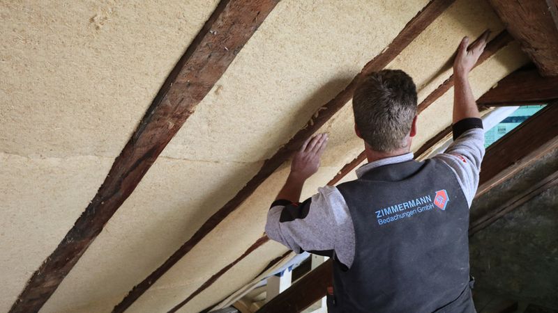 Ein Handwerker der Zimmermann Bedachungen GmbH montiert professionell eine ökologische Dachdämmung aus Holzfaserdämmplatten zwischen die Sparren eines Dachbodens.