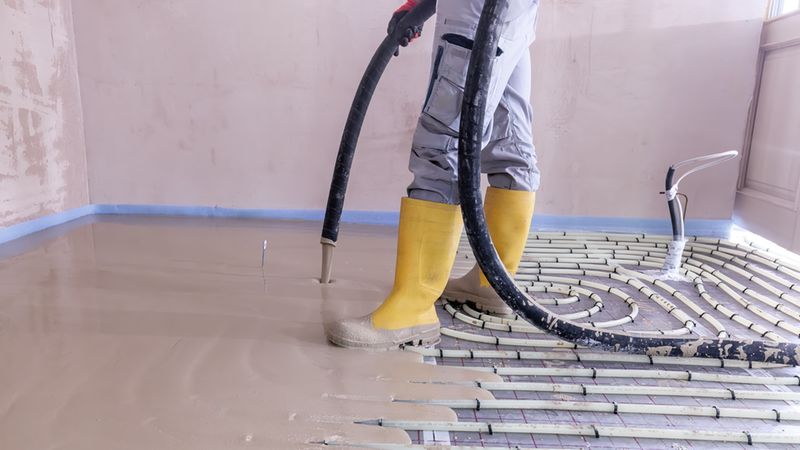 Ein Bauarbeiter in Schutzkleidung und gelben Gummistiefeln bringt mit einem Schlauch flüssigen Estrich über die bereits verlegten Rohre einer Fußbodenheizung auf einem Neubau-Boden ein.