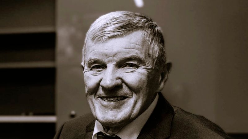 Ein professionelles Porträt eines älteren, freundlich lächelnden Herren mit grauen Haaren, der einen dunklen Anzug mit Krawatte trägt. Das Bild ist in einem sepia-artigen Stil gehalten.
