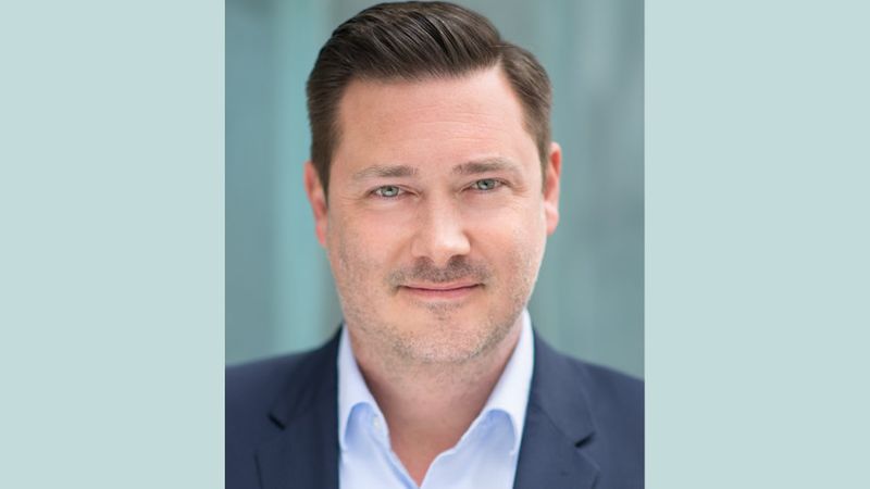 Ein professionelles Business-Porträt eines Mannes in den 40ern, der in ein hellblaues Hemd und ein dunkles Sakko gekleidet ist und freundlich in die Kamera lächelt. Der Hintergrund ist in einem dezenten, unscharfen Blauton gehalten.