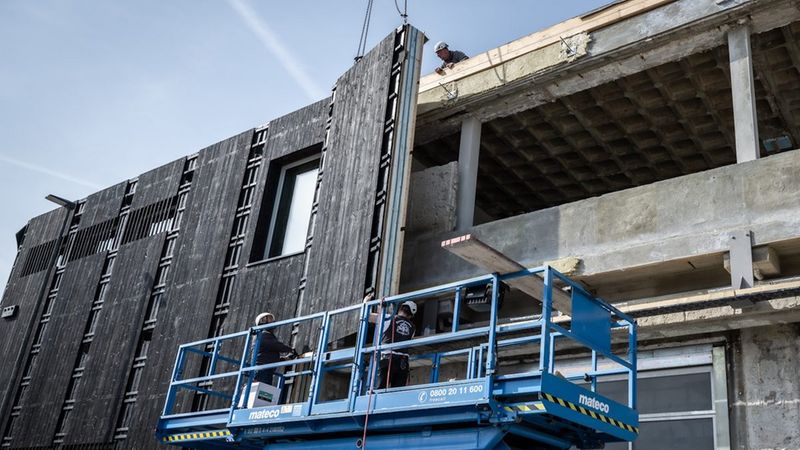 Bauarbeiter nutzen eine blaue Mateco-Hubarbeitsbühne, um eine dunkle Holzfassade an einem Industriegebäude zu montieren, während ein Kran das Fassadenelement hält.