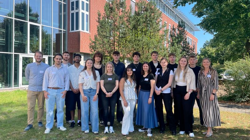 Eine große Gruppe von jungen Studierenden steht lächelnd vor einem modernen Universitätsgebäude mit roter Backsteinfassade und großen Glasfronten auf einer Grünfläche.