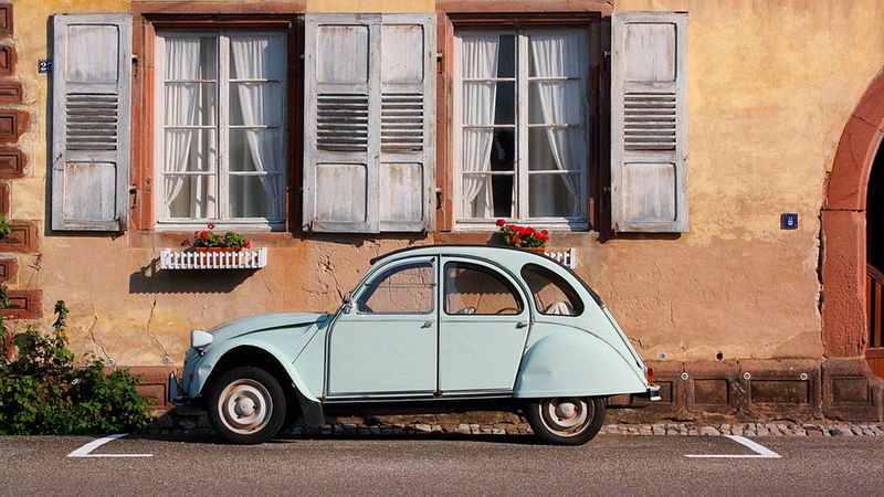 Ein hellblaues Citroen 2CV Auto (Ente) parkt vor einer charmanten, ockerfarbenen historischen Hausfassade mit weißen Holzfensterläden und kleinen Blumenkästen.
