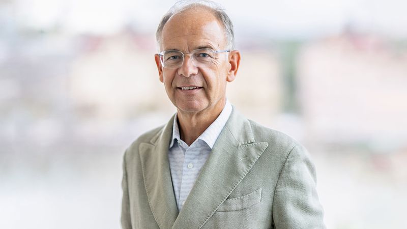 Ein älterer Mann in einem grauen Anzug steht vor einem verschwommenen Hintergrund und lächelt in die Kamera.