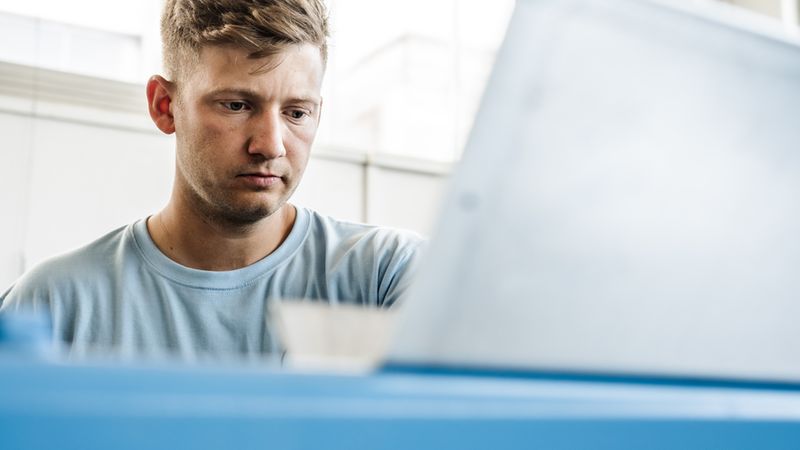 Ein junger Mann mit konzentriertem Gesichtsausdruck bedient eine industrielle Maschine in einer hellen Werkstattumgebung.
