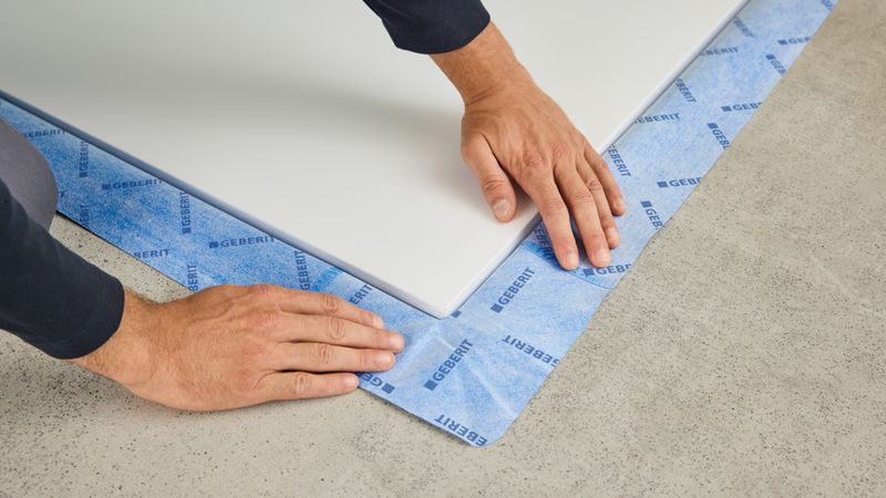 Ein Handwerker platziert eine weiße Duschwanne auf dem Boden, während er ein blaues Geberit Abdichtungsband fachgerecht an den Rändern anbringt, um eine sichere Abdichtung zu gewährleisten.