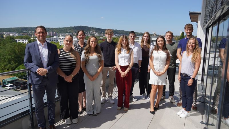 Eine Gruppe von jungen Auszubildenden und Mitarbeitern posiert gemeinsam mit ihrem Vorgesetzten auf einer sonnigen Dachterrasse für ein offizielles Teamfoto.