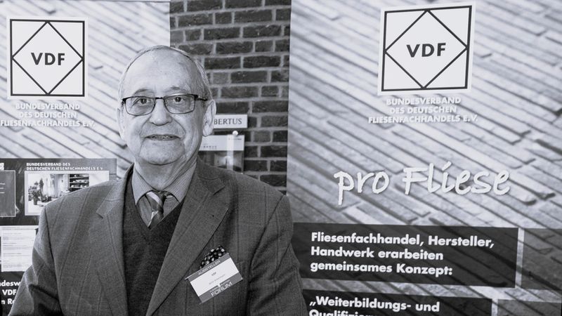 Ein älterer Herr im Anzug sitzt als Aussteller an einem Messestand des Bundesverbandes des Deutschen Fliesenfachhandels e.V. (VDF) vor einem Informationsbanner mit der Aufschrift 'pro Fliese'.
