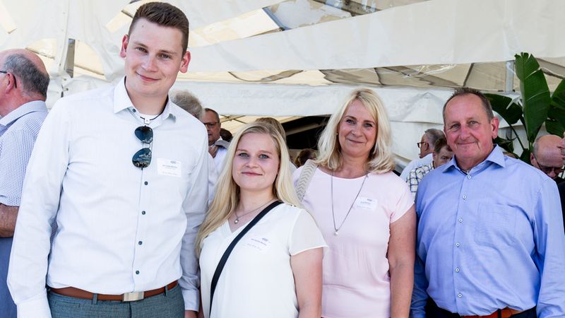 Eine Gruppe von vier Personen, bestehend aus zwei jüngeren Erwachsenen und zwei älteren Erwachsenen, die nebeneinander in einem hellen Festzelt für ein Foto posieren.