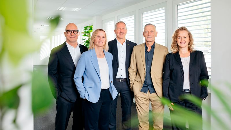 Ein professionelles Team von fünf Business-Experten, bestehend aus Männern und Frauen, steht in einem hellen, modernen Büroflur und lächelt freundlich in die Kamera.