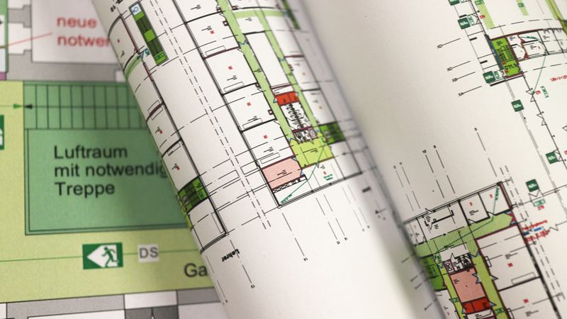 Ein detaillierter, aufgerollter Architektur-Grundrissplan auf weißem Papier, der die Raumaufteilung eines Gebäudes inklusive Treppenhäusern und Notausgang-Symbolen zeigt.