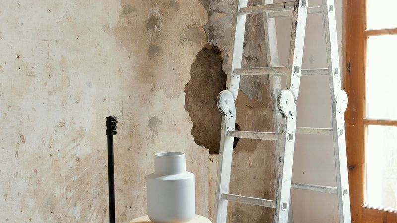 Eine Leiter und ein Hocker mit einer Vase stehen vor einer sanierungsbedürftigen Wand.