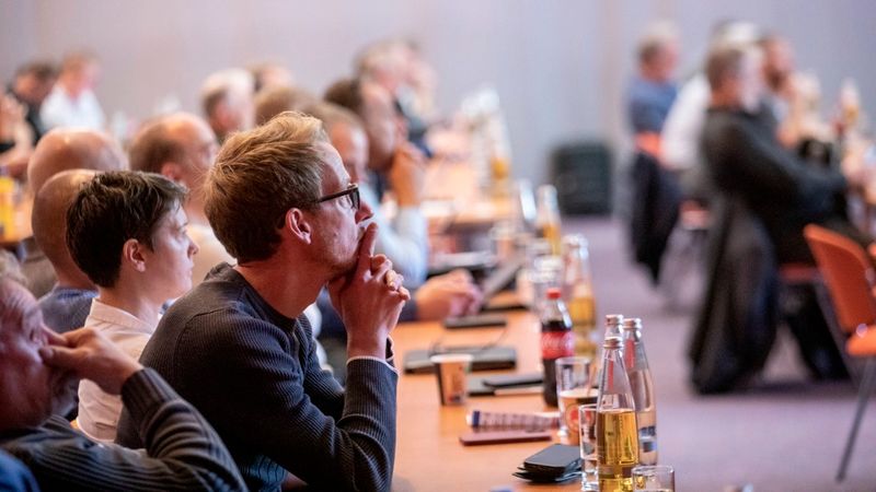 Eine Gruppe von aufmerksamen Konferenzteilnehmern sitzt an einem langen Tisch in einem Seminarraum und folgt konzentriert einem Vortrag.