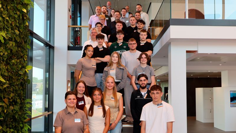 Ein großes Gruppenfoto von Auszubildenden und Mitarbeitern, die auf einer breiten Treppe in einem modernen Bürogebäude posieren. Die Gruppe ist nach oben hin gestaffelt und zeigt eine vielfältige Mischung von jungen Erwachsenen und erfahrenen Kollegen vor einer begrünten Wand.