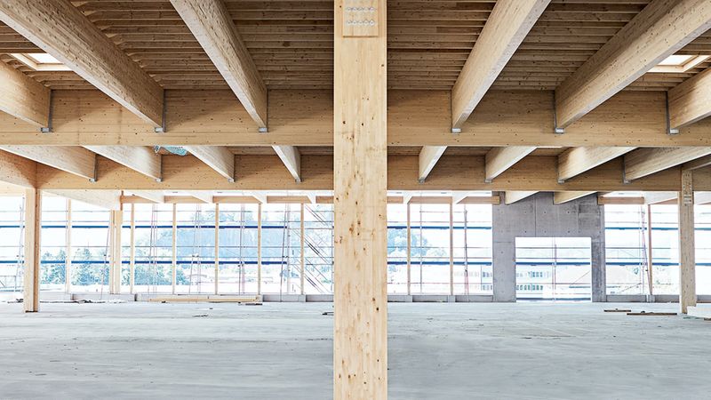 Innenansicht einer modernen Baustelle mit einer beeindruckenden Holzkonstruktion, die eine weitläufige Hallenfläche überspannt, während im Hintergrund eine Fensterfront und Betonelemente erkennbar sind.