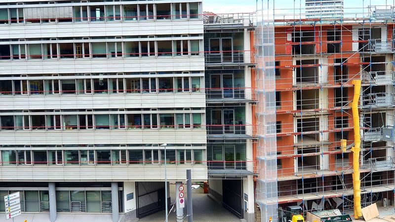 Eine Außenansicht eines Gebäudekomplexes, der aus einem modernen Bürogebäude und einem angrenzenden, eingerüsteten Wohnhaus besteht. Das Wohnhaus befindet sich derzeit in der Sanierung, was durch das umfangreiche Baugerüst und die offene Fassade deutlich wird.