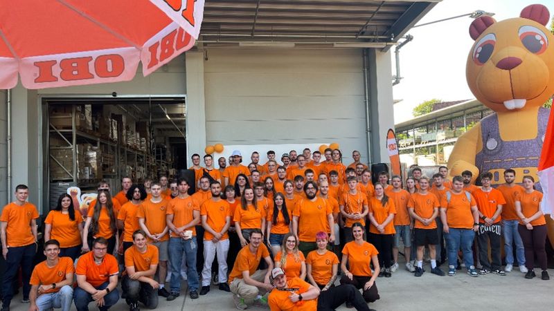 Ein großes Team von Mitarbeitern in orangefarbenen T-Shirts versammelt sich für ein Gruppenfoto vor einem OBI-Baumarkt, begleitet von einem großen aufblasbaren OBI-Biber.