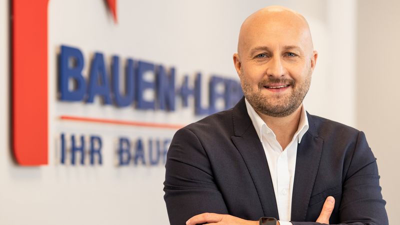 Ein professionelles Porträt eines freundlich lächelnden Unternehmers im dunklen Anzug, der mit verschränkten Armen vor einem Firmenlogo von 'Bauen + Leben' steht.