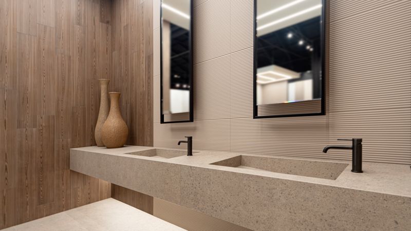 Ein stilvolles, modernes Doppelwaschbecken in einem minimalistisch gestalteten Badezimmer. Die Waschtischeinheit besteht aus einem sandfarbenen Steinmaterial mit zwei integrierten Becken und schwarzen Armaturen. An der Wand hängen zwei schmale, vertikale Spiegel über den Waschbecken, ergänzt durch eine Kombination aus holzoptik-vertäfelten Wänden und strukturierten hellen Fliesen sowie zwei dekorativen Vasen.