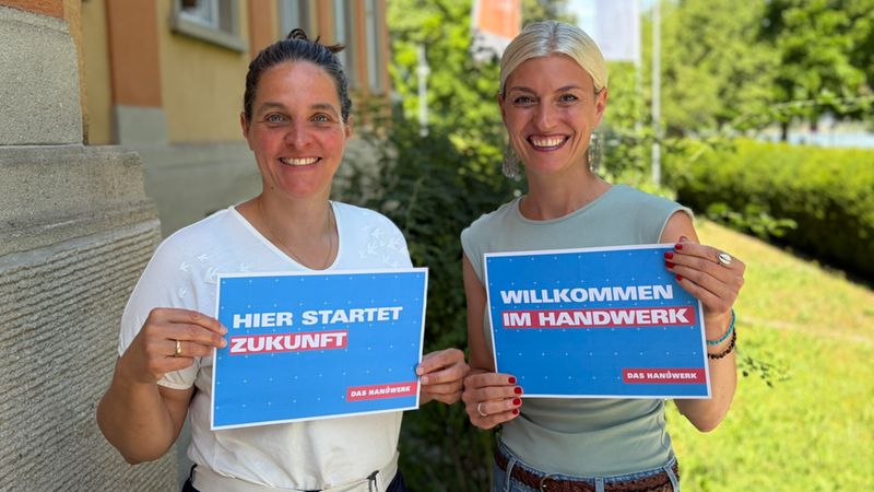 Zwei lächelnde Frauen halten Werbeschilder der Kampagne 'Das Handwerk' in den Händen, mit den Aufschriften 'Hier startet Zukunft' und 'Willkommen im Handwerk', vor einer Außenkulisse.