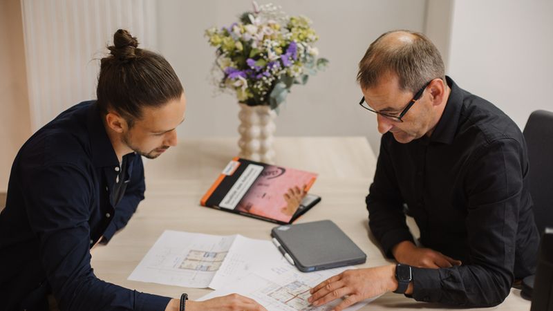 Ein junger Mann und ein erfahrener Architekt sitzen an einem hellen Holztisch und besprechen konzentriert detaillierte Baupläne. Auf dem Tisch liegen neben den Entwürfen ein Tablet und eine dekorative Blumenvase, was die professionelle und dennoch einladende Arbeitsatmosphäre unterstreicht.