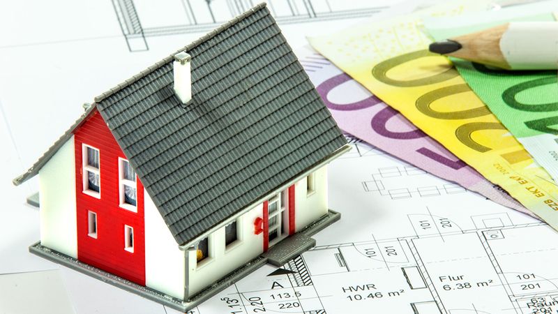 Ein Modellhaus steht auf einem  Bauplan, umgeben von Euro-Geldscheinen und einem Bleistift.