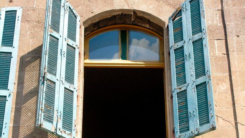 Ein detaillierter Blick auf ein historisches Steinhaus mit einem bogenförmigen Fenster, das von verwitterten, hellblauen Fensterläden aus Holz eingerahmt wird.