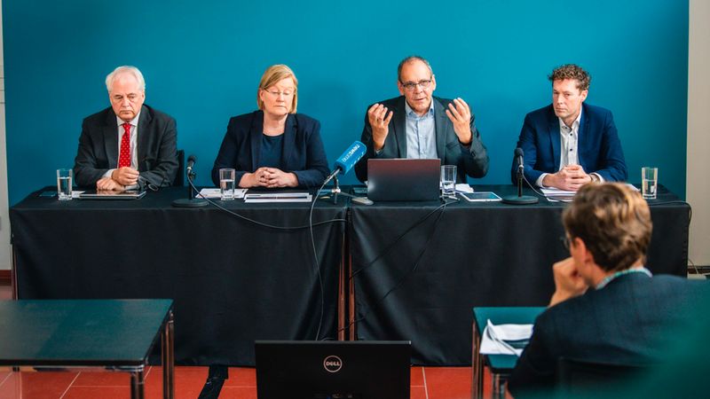 Eine Gruppe von vier Experten sitzt hinter einem mit schwarzem Stoff bedeckten Konferenztisch bei einer Pressekonferenz vor einer türkisfarbenen Wand. Der Mann in der Mitte spricht aktiv und gestikuliert mit seinen Händen, während die anderen drei aufmerksam zuhören oder sich Notizen machen. Auf dem Tisch stehen Mikrofone, Laptops und Gläser.