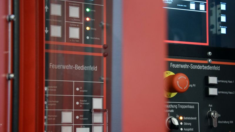 Nahaufnahme eines modernen Feuerwehr-Bedienfeldes und eines Feuerwehr-Sonderbedienfeldes mit Not-Aus-Schalter, Anzeigelampen und verschiedenen Tastern zur Steuerung von Brandmelde- und Entrauchungsanlagen.