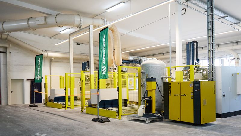 Helle Industriehalle mit einer auffälligen gelben Kompressorstation und Druckluftanlage als Hauptmotiv. Sichtbar sind mehrere gelbe Maschinen, ein großer Druckbehälter, Rohrleitungen und Trenngitter in einer sauberen Produktionshalle. Die Szene zeigt technische Anlagen und Fördertechnik in einer modernen Betriebsumgebung.