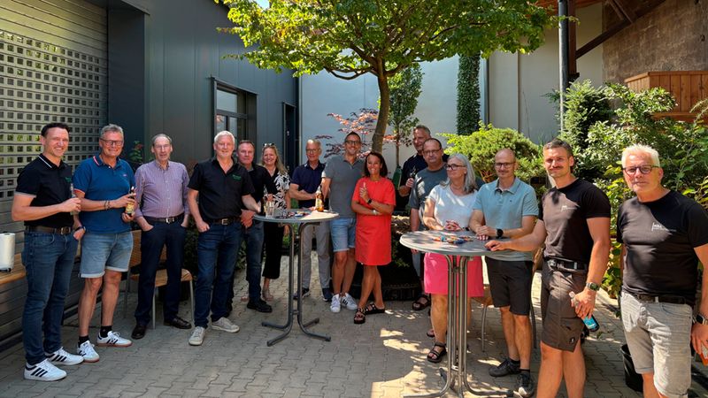 Eine Gruppe von Mitarbeitern steht entspannt zusammen bei einem Firmenevent in einem sonnigen Innenhof, umgeben von Pflanzen und Stehtischen.