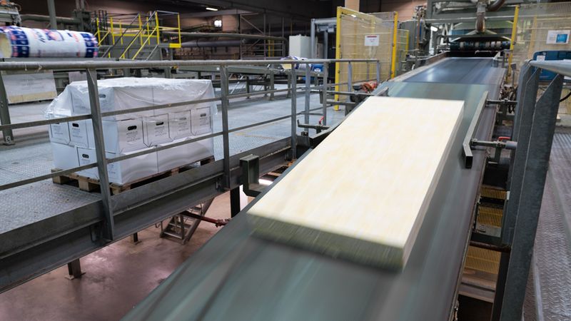 Eine industrielle Produktionsanlage für Dämmstoffe, bei der eine Dämmplatte auf einem Förderband transportiert wird, während im Hintergrund palettierte Pakete lagern.